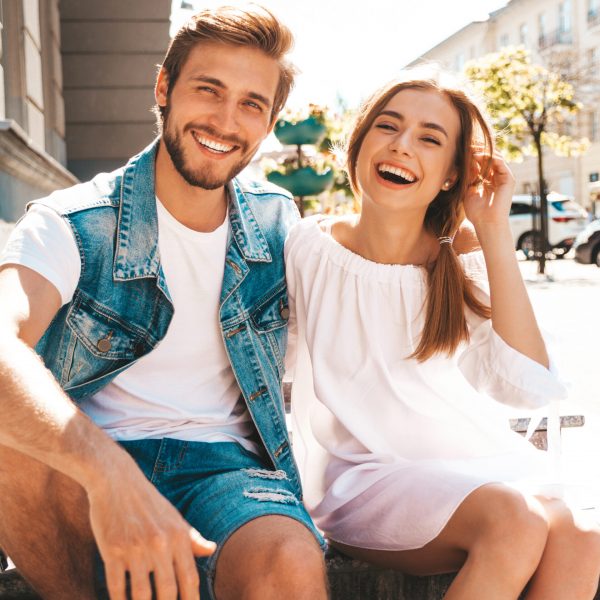 Smiling beautiful girl and her handsome boyfriend. Woman in casual summer dress and man in jeans clothes. Happy cheerful family. Sitting on stairs on the street background.Hugging couple